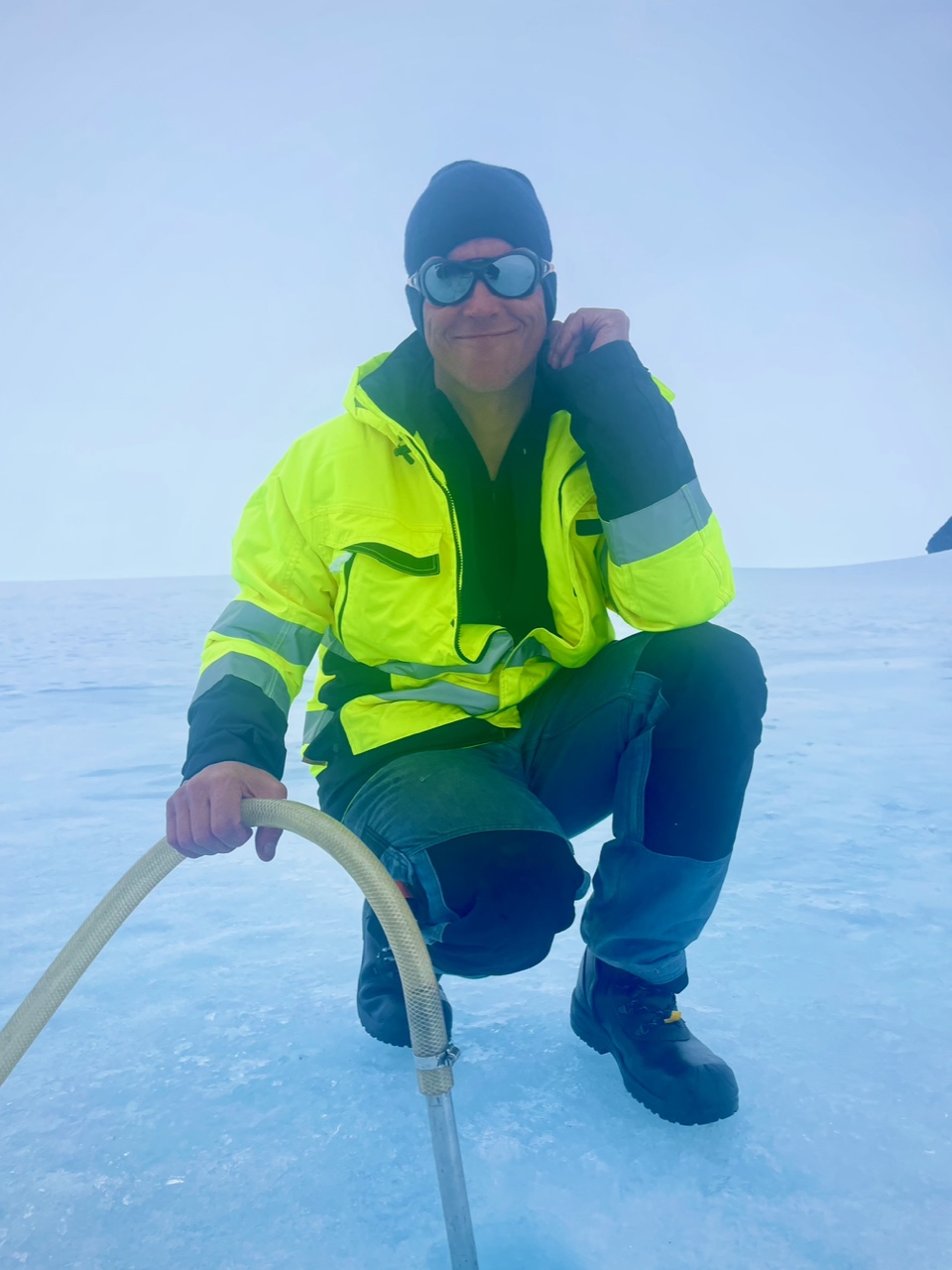 En man i gul jacka och solglasögon sitter hukad på isen.