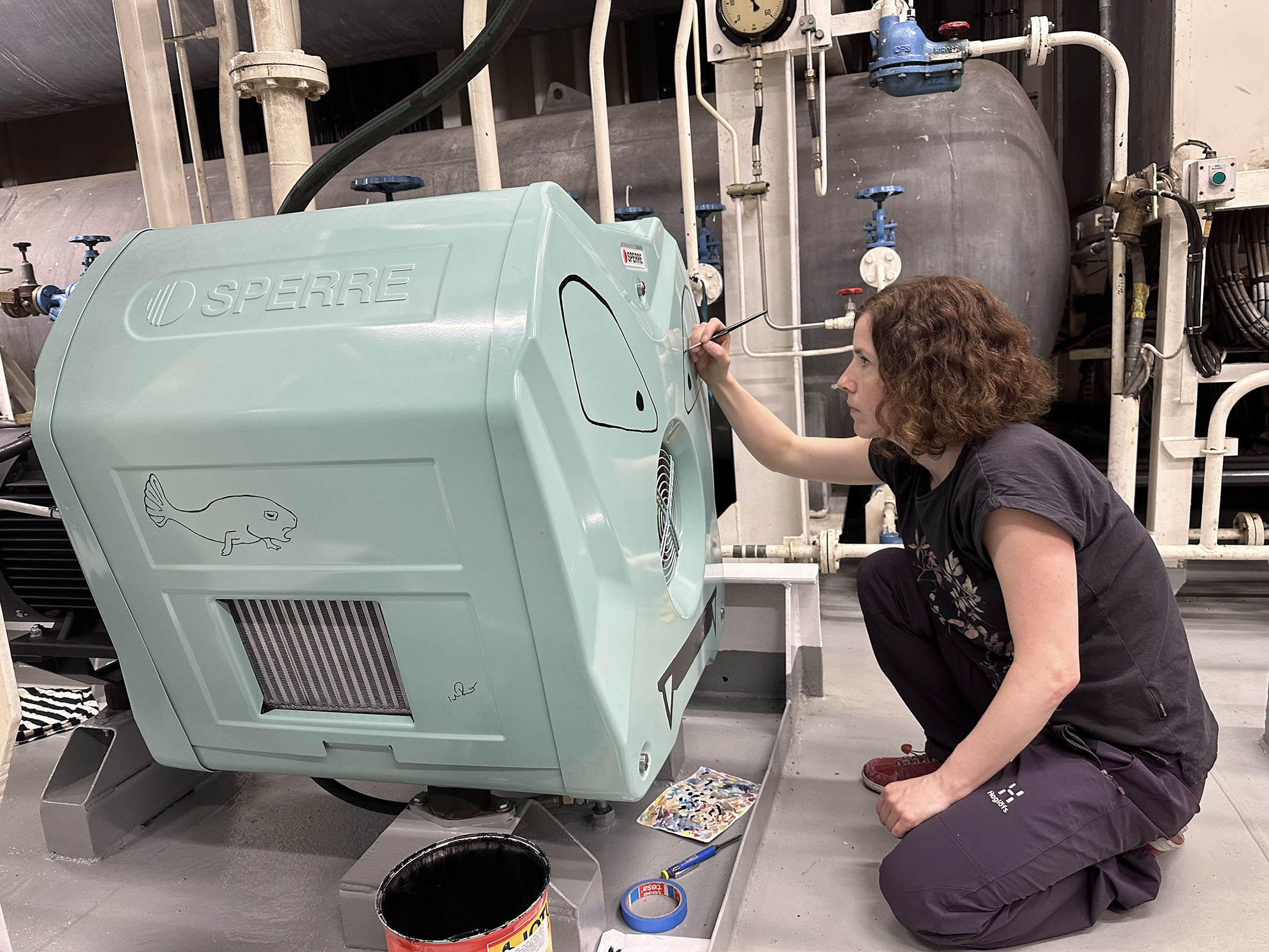 En mintgrön startluftskompressorn målas med ögon, mun och vassa tänder. Konstnären sitter på huk med en pensel. 