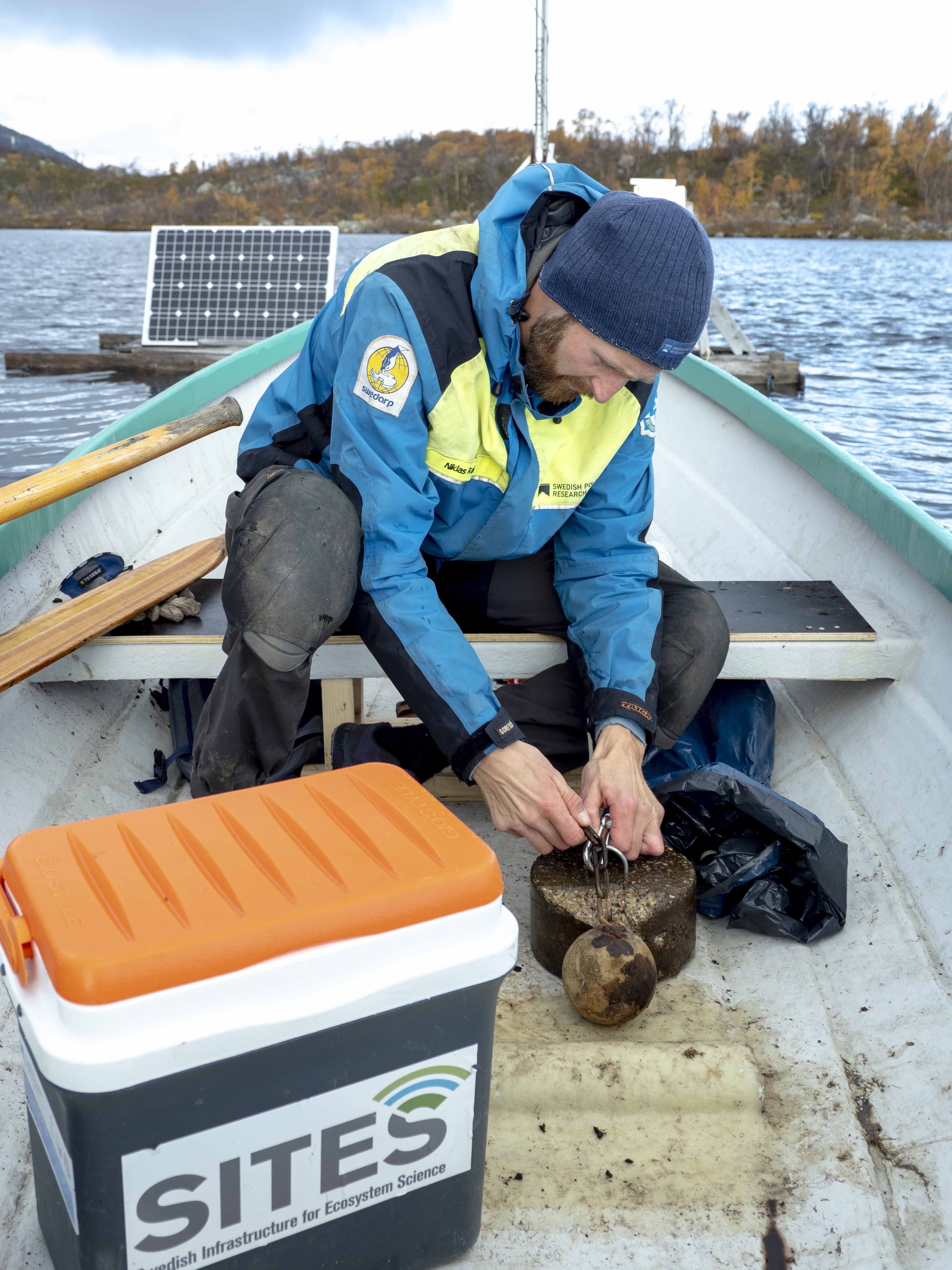 En man i blå och gul jacka sitter i en båt böjd över en metallvikt