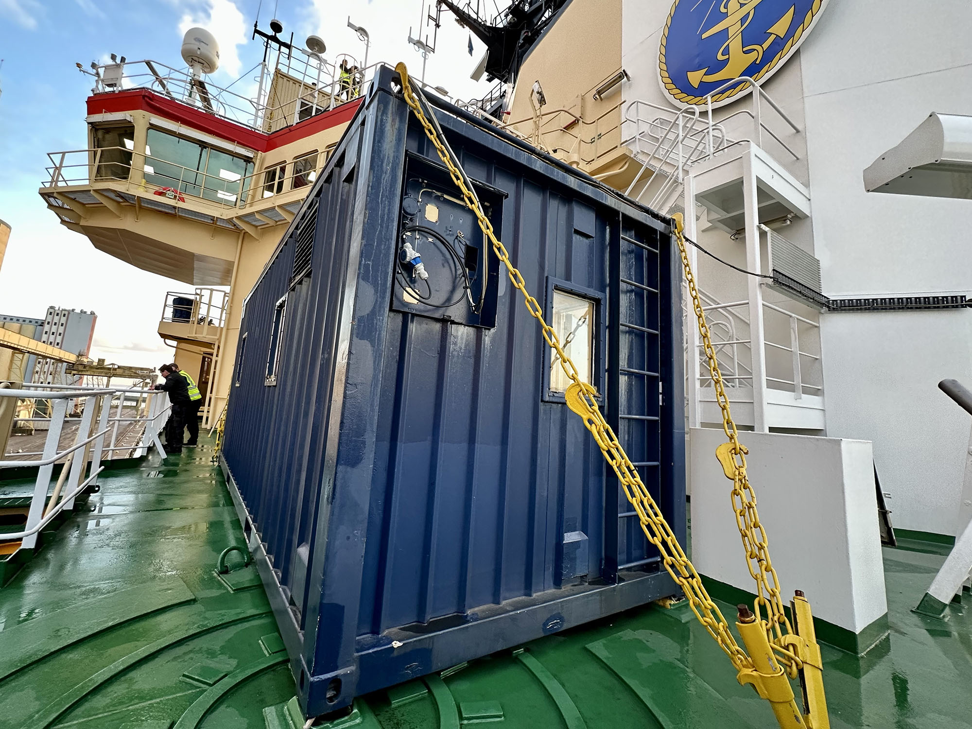 En blå container står stadigt förankrad med gula kättingar i det gröna fartygsdäcket. 